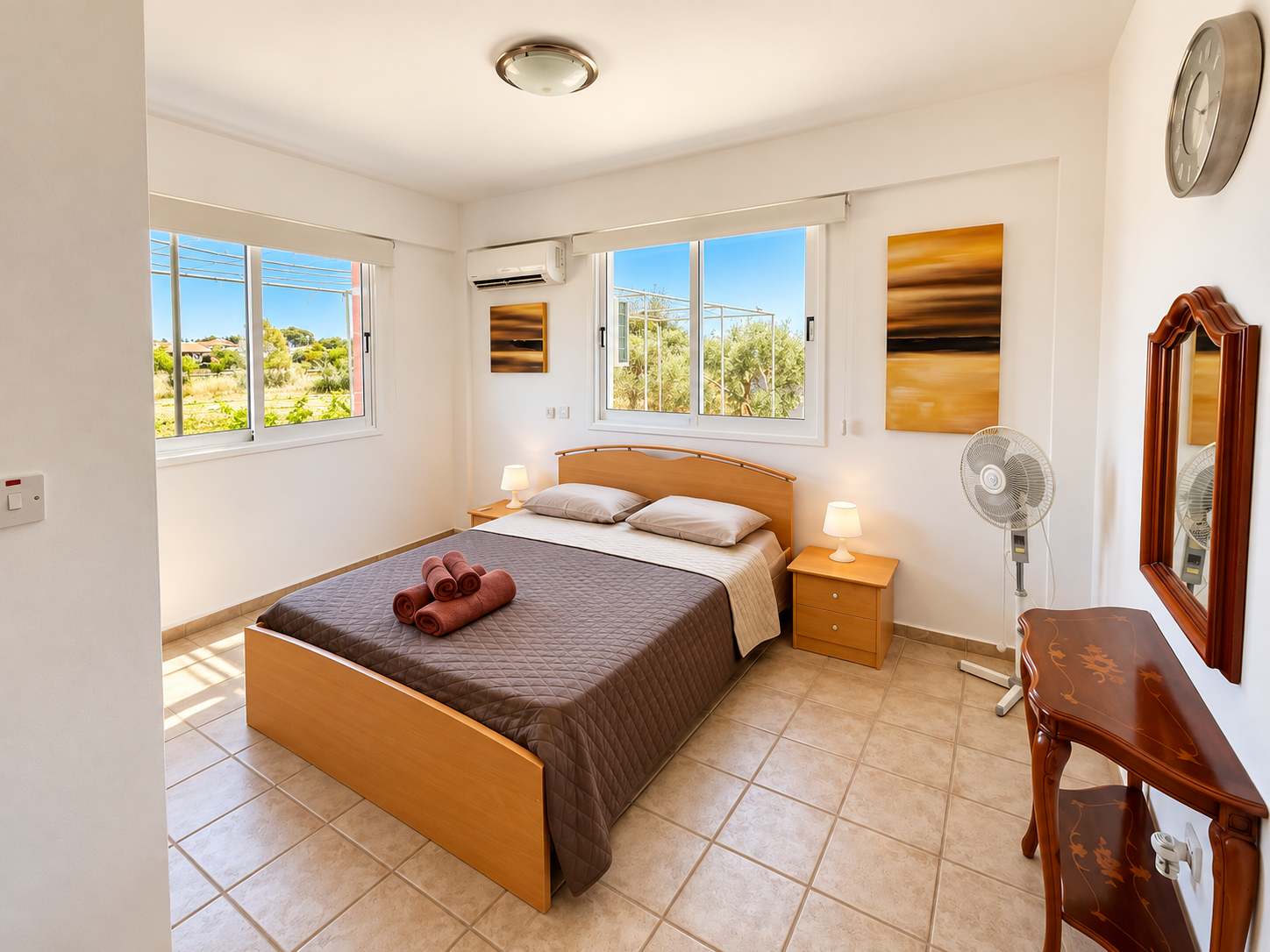 A made bed beneath a ceiling fan, with greenery visible through the windows
