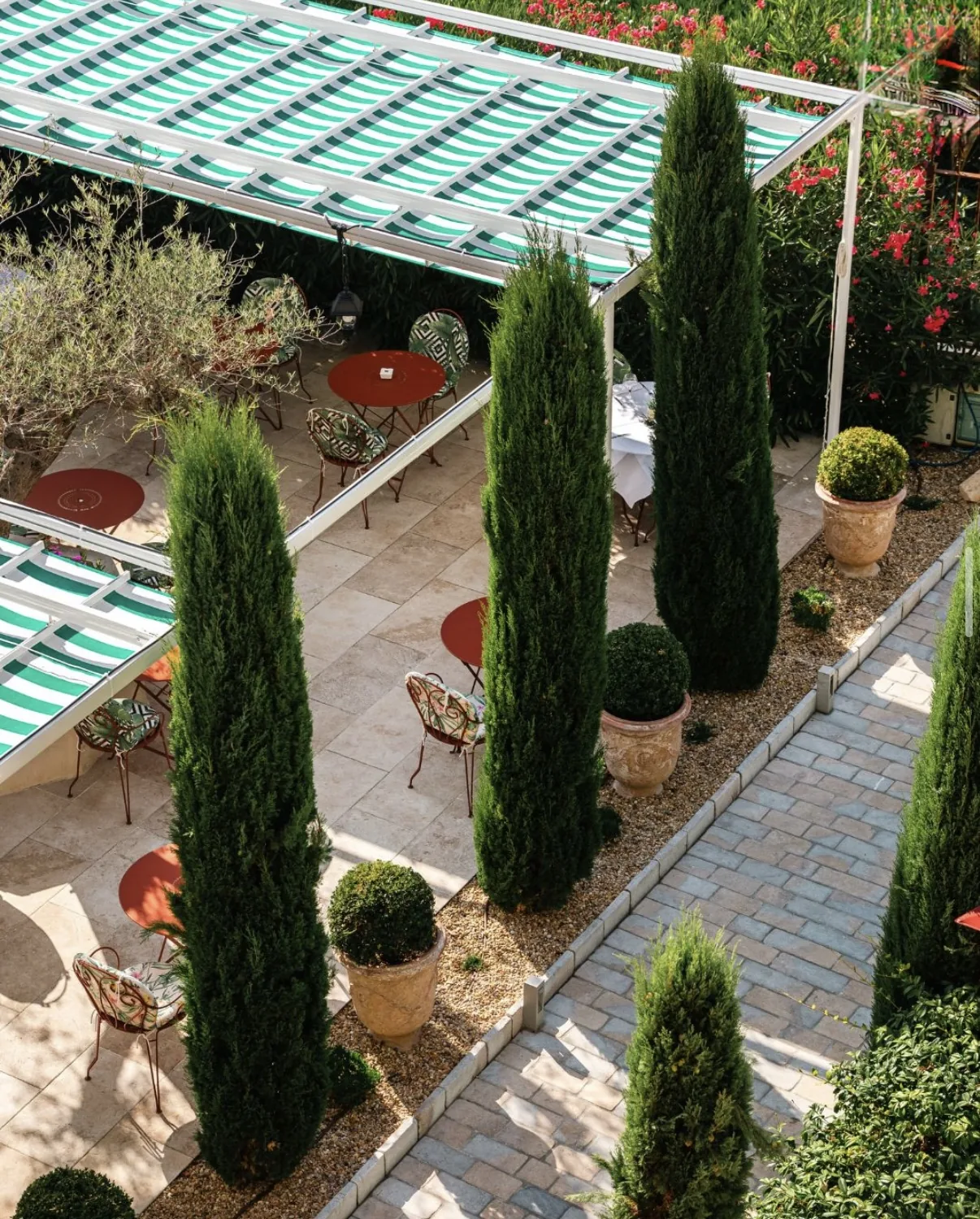 La terrasse vue d'en haut : stores rayés verts, tables rouges et cyprès du jardin